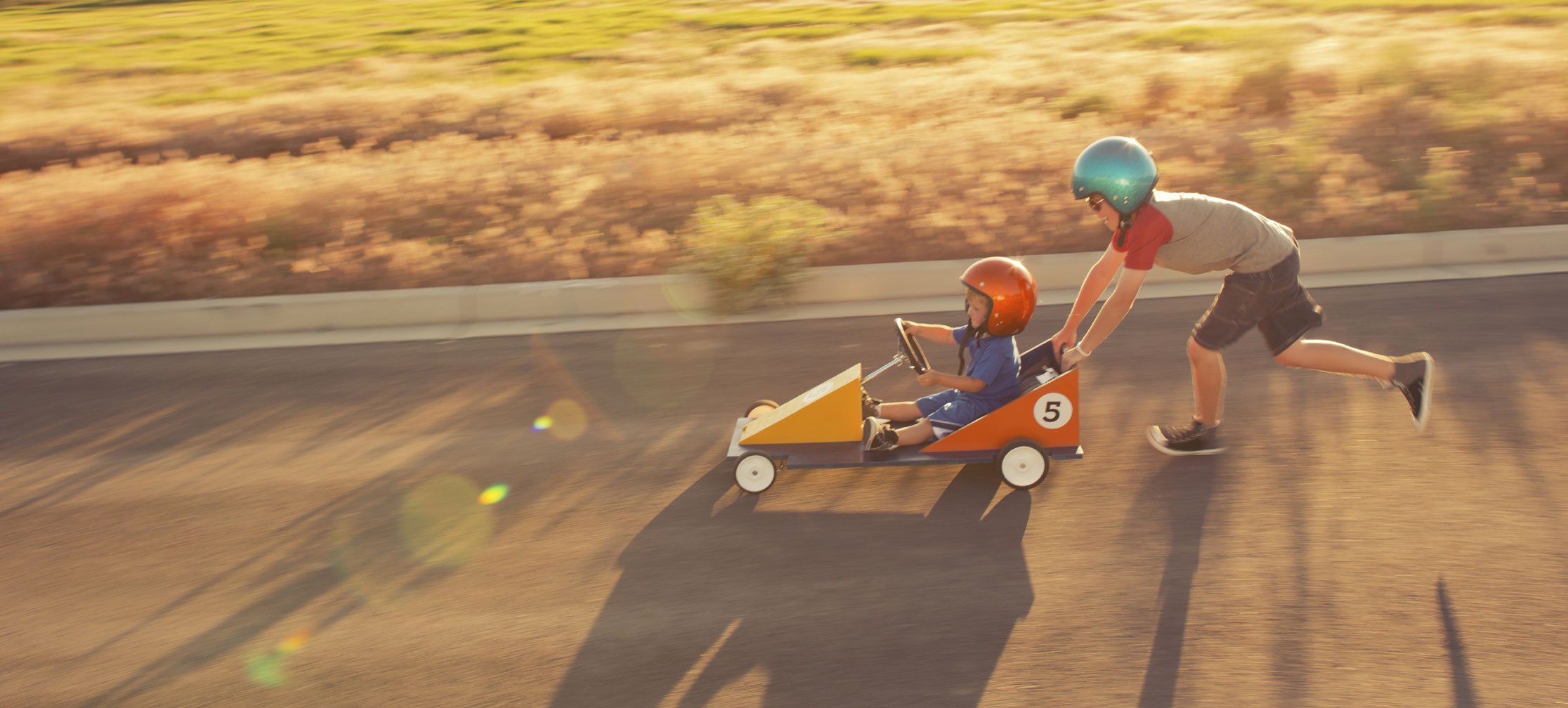 Image of a child pushing another child down the road on a billycart.