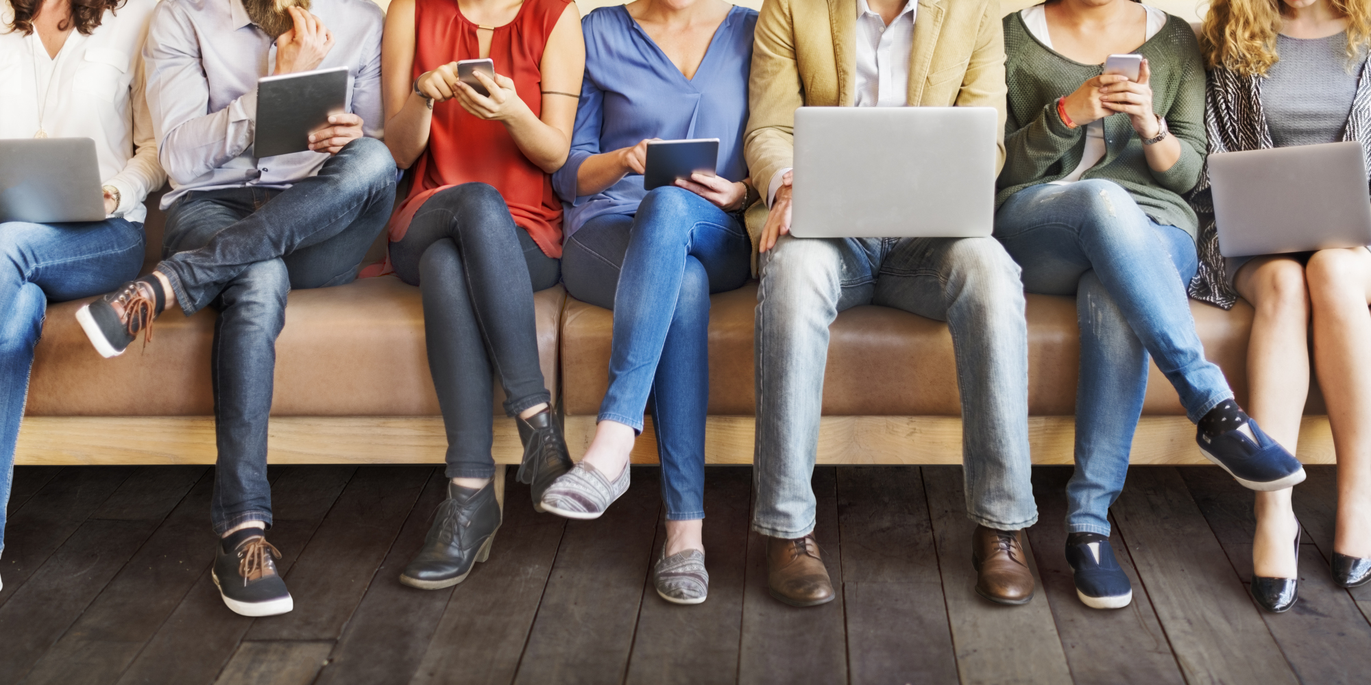 Image showing a row of people sitting on a couch with their legs crossed looking at different devices.