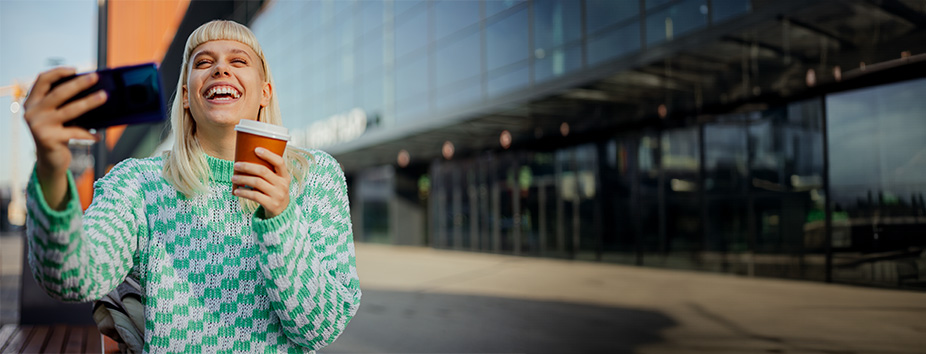 A young woman with light skin tone is laughing while having a video call on the street. She's holding her mobile phone in one hand and a takeaway coffee cup in the other hand. 