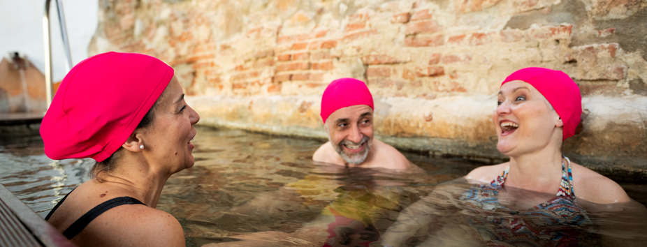 Three people with a range of skin tones and genders are sitting in an outdoor pool. They are all wearing pink swimming caps and are smiling and talking to each other.