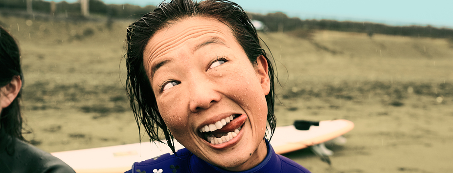 woman with medium skin tone is smiling and sticking her tongue out, glancing sideways. She’s at the beach and is wearing a wetsuit, with a surfboard in the background. 