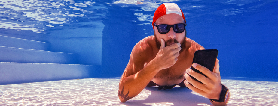 A man with medium skin tone and a beard is lying at the bottom of a pool looking at his phone. He’s wearing a swimming cap and sunglasses.