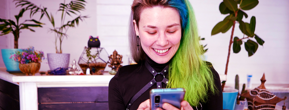 A young woman with light skin tone is holding her phone with two hands, looking down at it smiling. She has green, blue and pink hair. 