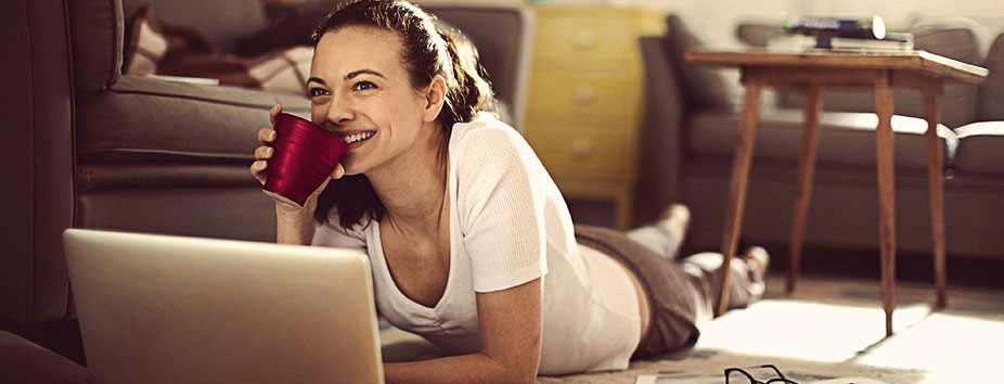 A woman drinking a cup of coffee, smiling, looking at a budget spreadsheet on her laptop