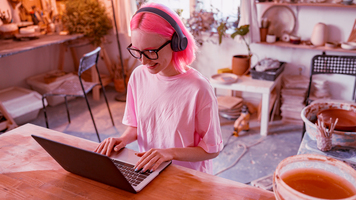 High angle view of happy woman artisan wearing wireless headphones sitting at table and working with laptop in ceramics workshop