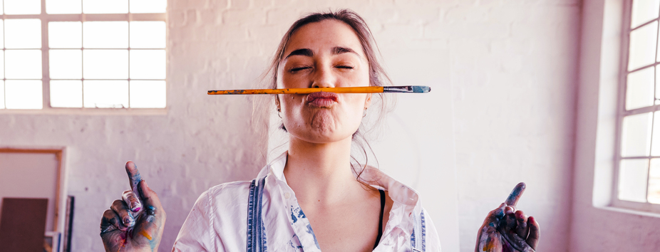 A woman with light skin tone is wearing overalls. She has paint on her hands, her eyes are closed and is facing the camera. She’s balancing a paintbrush above her upper lip.