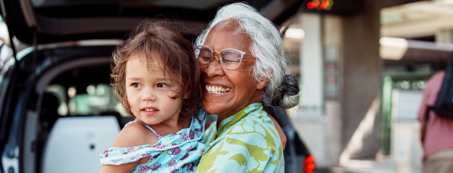 An older woman with dark skin tone is smiling and standing, holding a child with medium skin tone. There’s a car with the boot open in the background. 