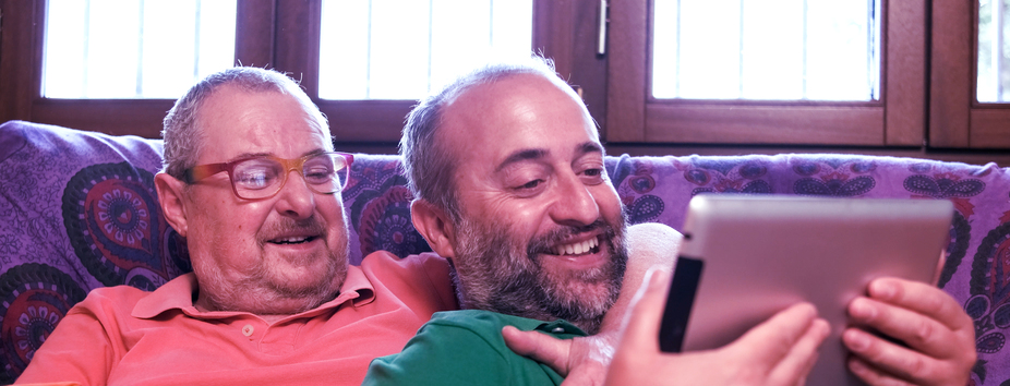 Two men with medium skin tone are lying on a couch. One man is embracing the other, and they’re both smiling looking at a tablet device. 