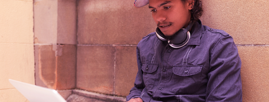 A young man with dark skin tone is sitting on the ground with his back against a wall. He’s typing with both hands on a laptop. 