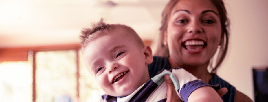 A woman with medium skin tone is holding her toddler. They’re both facing the camera and laughing. 