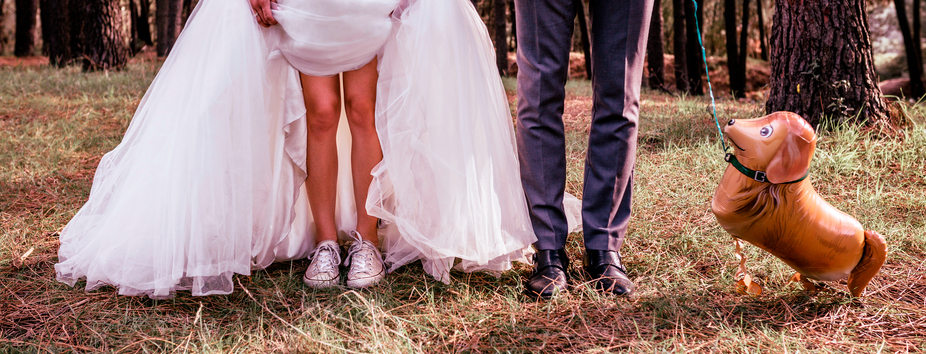 A couple with light skin tone are seen from the torso down. They’re facing the camera. The woman is wearing a wedding dress which is held up to show white sneakers. The man is holding a dog shaped balloon.