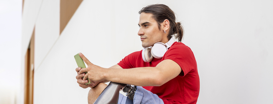 A man with light skin tone and long hair is sitting on a skateboard holding his phone with two hands. His left leg is prosthetic. 