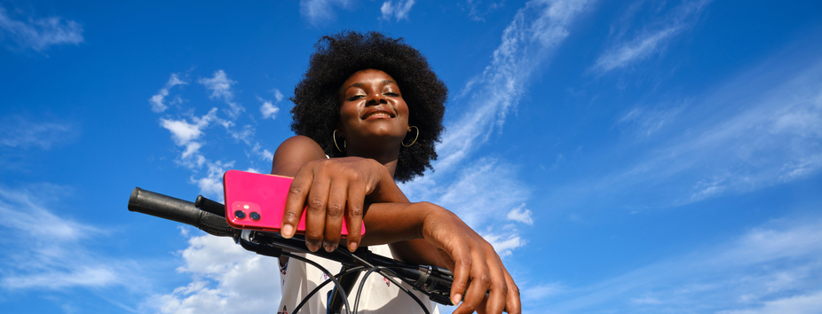 A young woman with dark skin tone is leaning over a bicycle, holding her phone in one hand. She’s smiling at the camera.