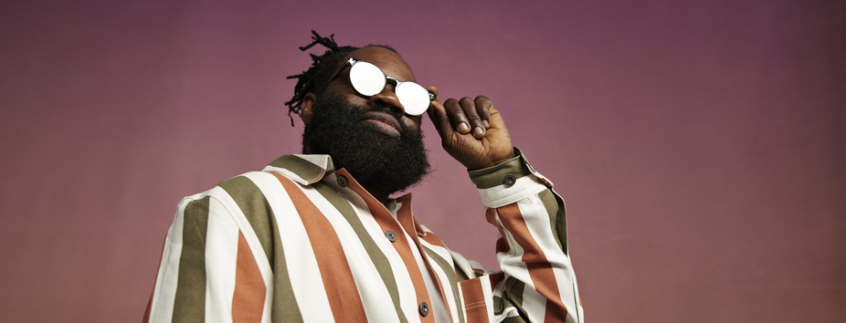 A man with dark skin tone, dreadlocks and a beard is wearing sunglasses and looking down at the camera.