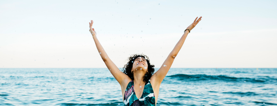 A woman with medium skin tone is standing in the ocean with her arms outstretched and head looking up. She’s wearing a one-piece swimming costume.